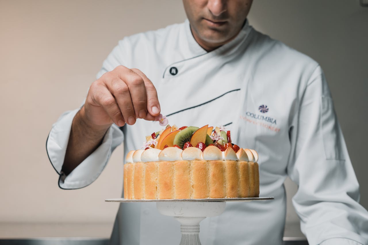 A pastry chef in a white coat delicately places an edible flower on a fruit-topped charlotte cake, which is decorated with whipped cream, strawberries, kiwi, and peaches.