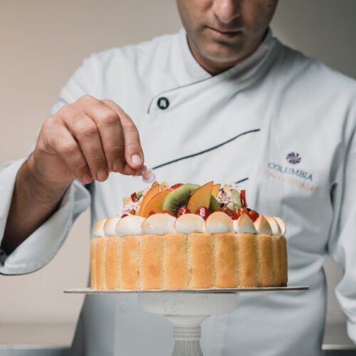A pastry chef in a white coat delicately places an edible flower on a fruit-topped charlotte cake, which is decorated with whipped cream, strawberries, kiwi, and peaches.