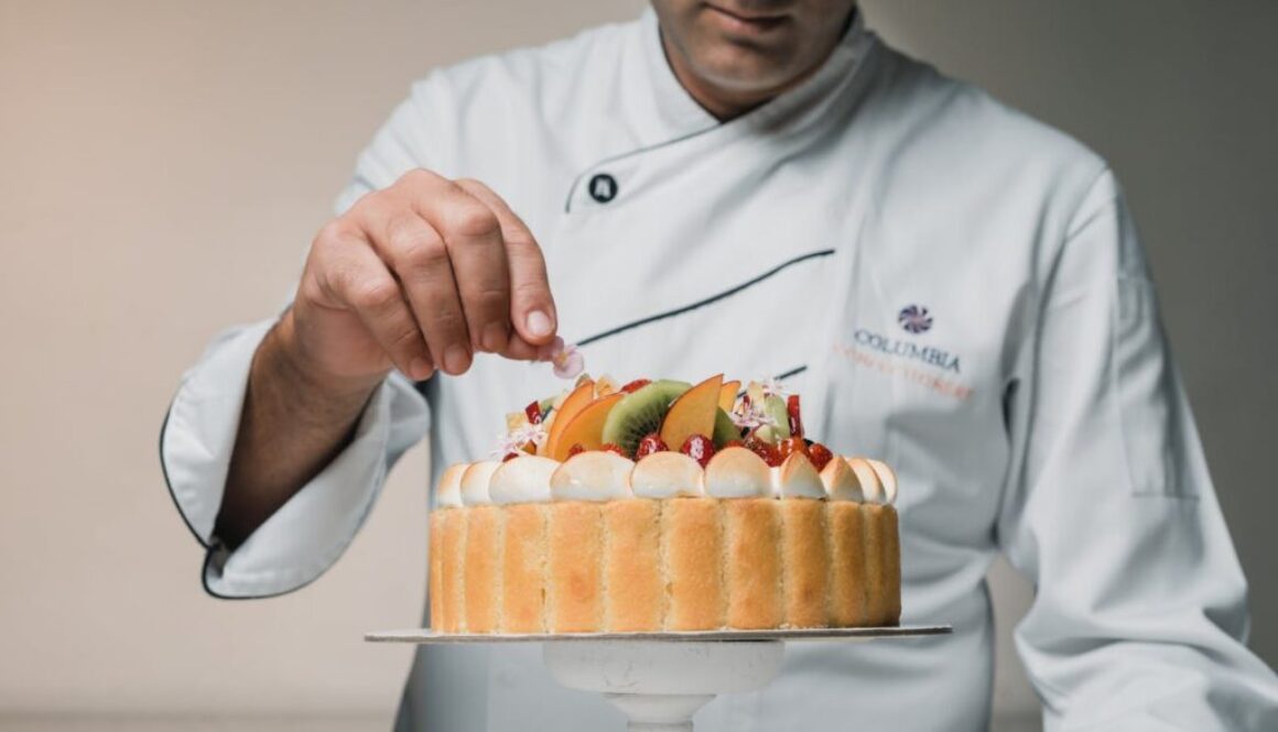 A pastry chef in a white coat delicately places an edible flower on a fruit-topped charlotte cake, which is decorated with whipped cream, strawberries, kiwi, and peaches.